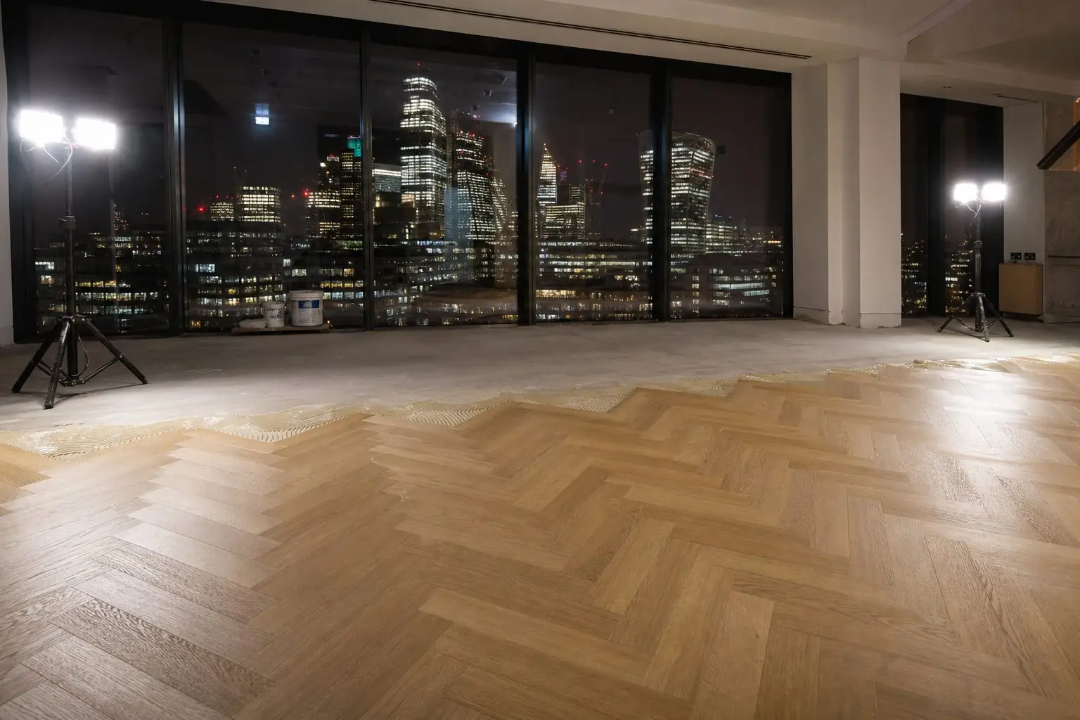 Oak herringbone floor being laid overnight in a London office, trade work lights on and the city skyline lit up through the windows